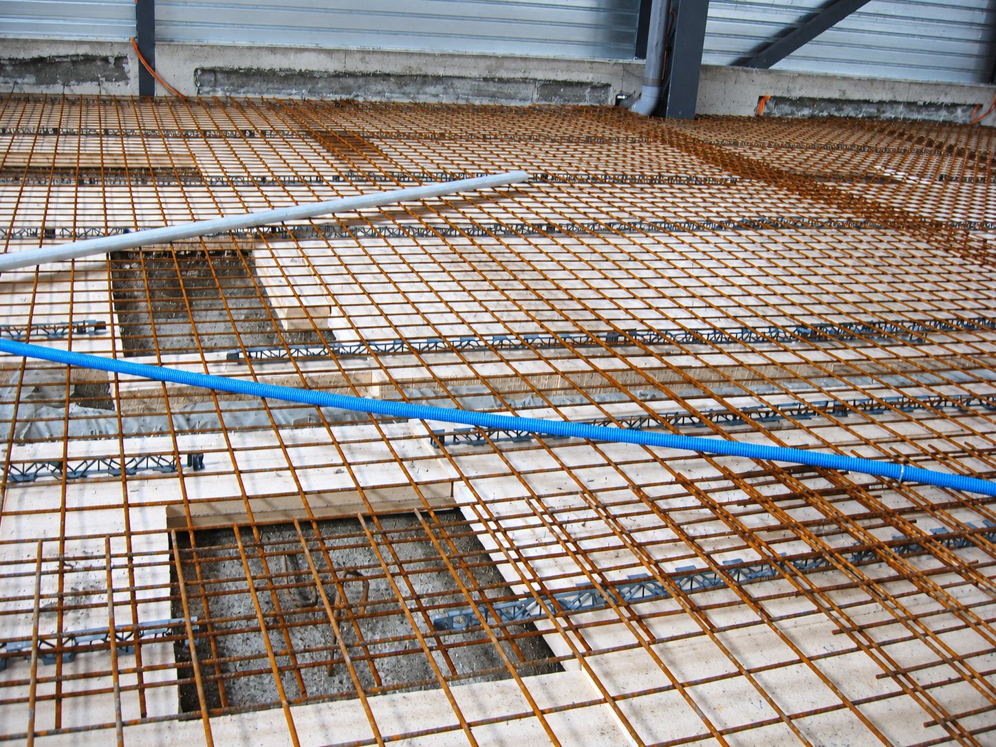 Vue d'un chantier montrant un treillis soudé rouillé posé sur des panneaux isolants blancs, avec une gaine bleue et un tube gris en travers.