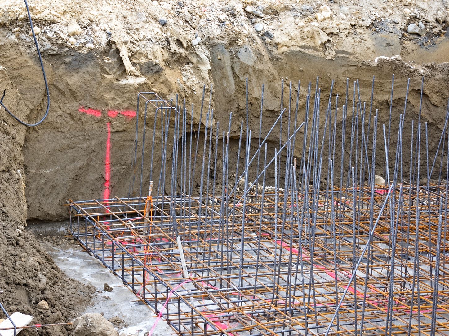 Un treillis d'armatures en acier avec des tiges verticales au fond d'une excavation, devant une paroi de terre marquée de peinture rose.
