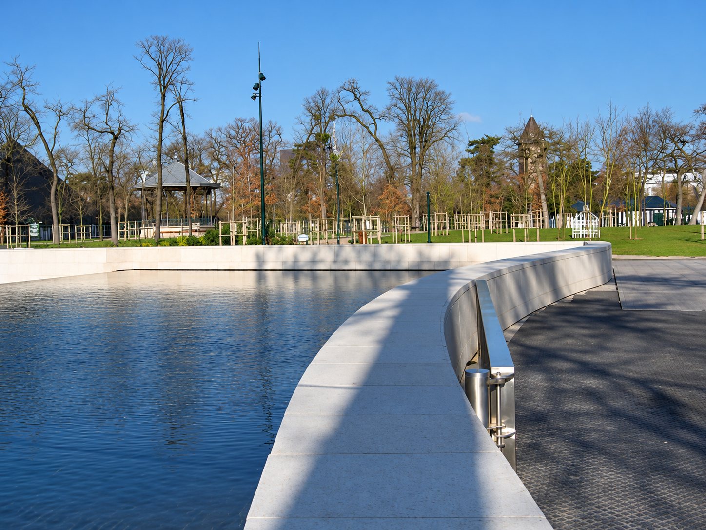 Un grand bassin d'eau calme aux bords courbes en pierre claire avec une rambarde en inox, entouré d'arbres sans feuilles dans un parc.