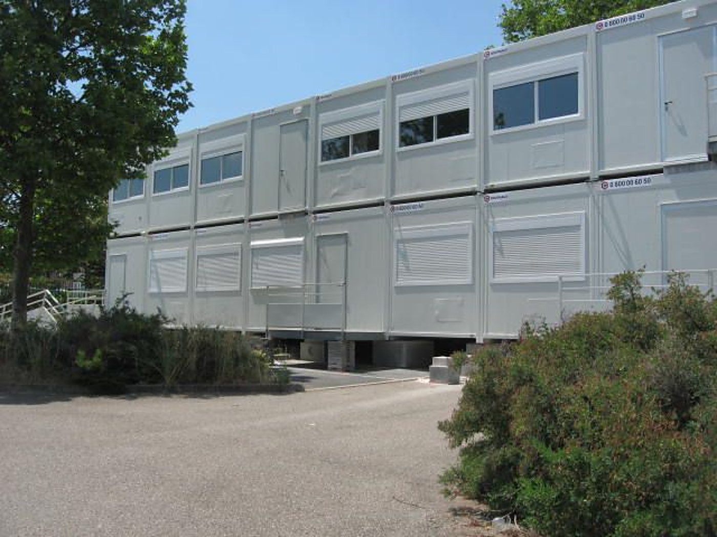 Bâtiment modulaire gris à deux niveaux, fait d'éléments juxtaposés avec fenêtres et volets roulants, entouré d'arbustes et d'un parking en gravier.