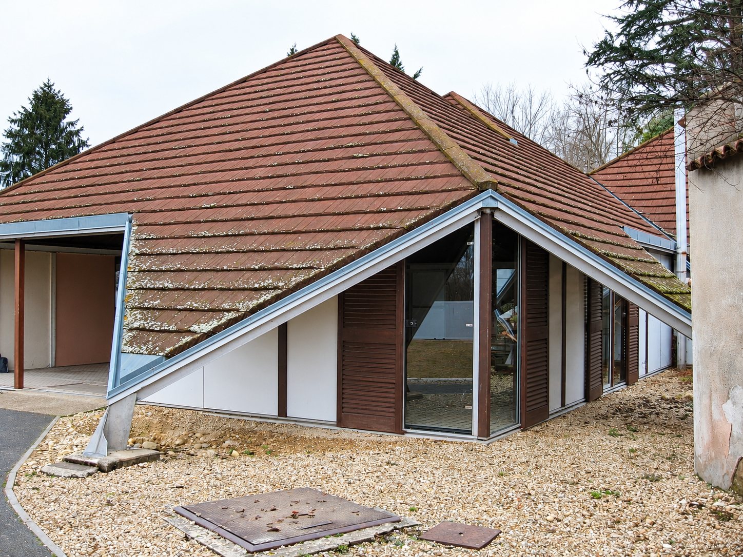 Un bâtiment de plain-pied au grand toit pyramidal en tuiles brunes mousseuses, avec une façade vitrée, des volets bruns et un sol de gravier.