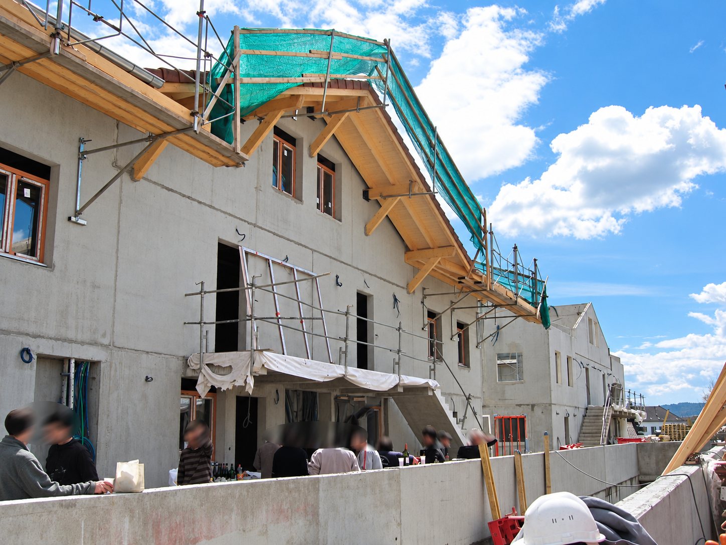 Façade en béton d'un bâtiment en construction avec charpente en bois, filet vert et échafaudages, tandis que des personnes se tiennent sur une terrasse au premier plan.