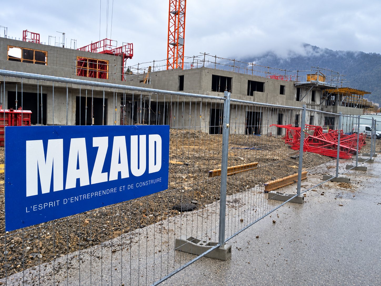 Un chantier de construction de bâtiments en béton, entouré d'une clôture grillagée portant un panneau bleu Mazaud, sous un ciel brumeux avec une grue en arrière-plan.