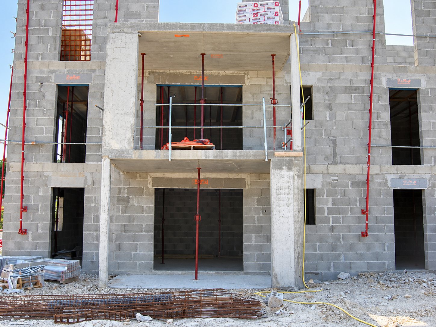 Façade d'un bâtiment en cours de construction, en blocs de béton, avec des étais métalliques rouges, des ouvertures vides et des armatures posées au sol.