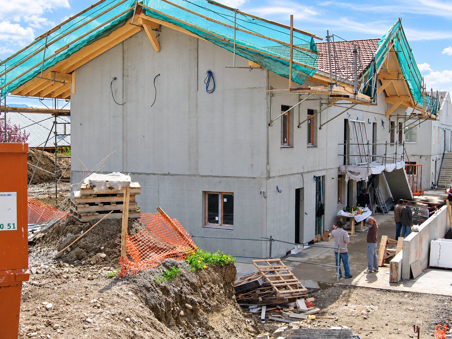 Maison en béton brut en construction, avec charpente bâchée, échafaudages, terre excavée et ouvriers sur le chantier.
