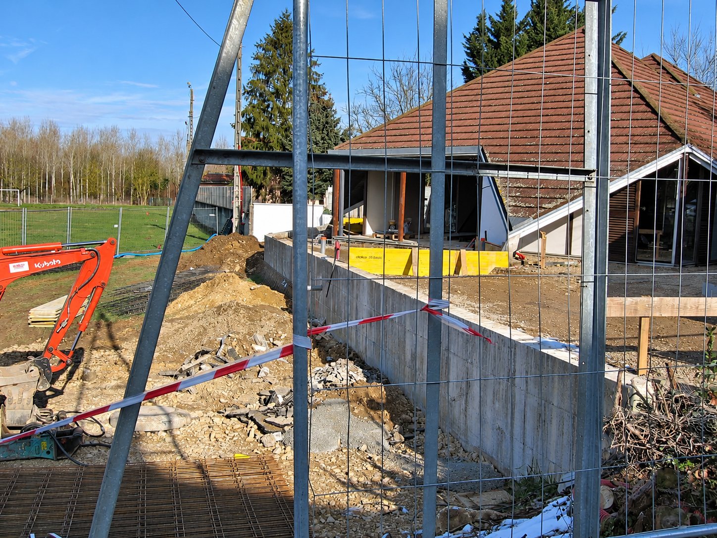 Vue à travers une clôture de chantier sur un mur en béton, des terres excavées, une pelleteuse orange et un bâtiment à toit de tuiles.