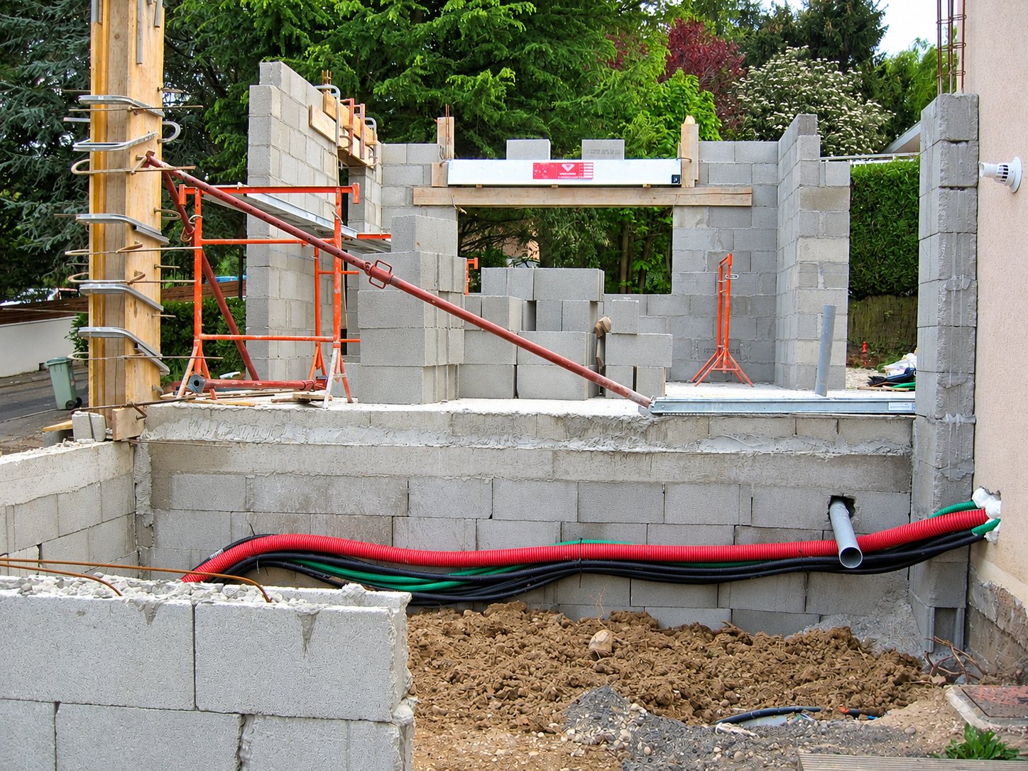 Chantier de maison en blocs de béton avec échafaudage orange, gaines rouges et noires posées sur le mur et terre fraîchement remuée au premier plan.