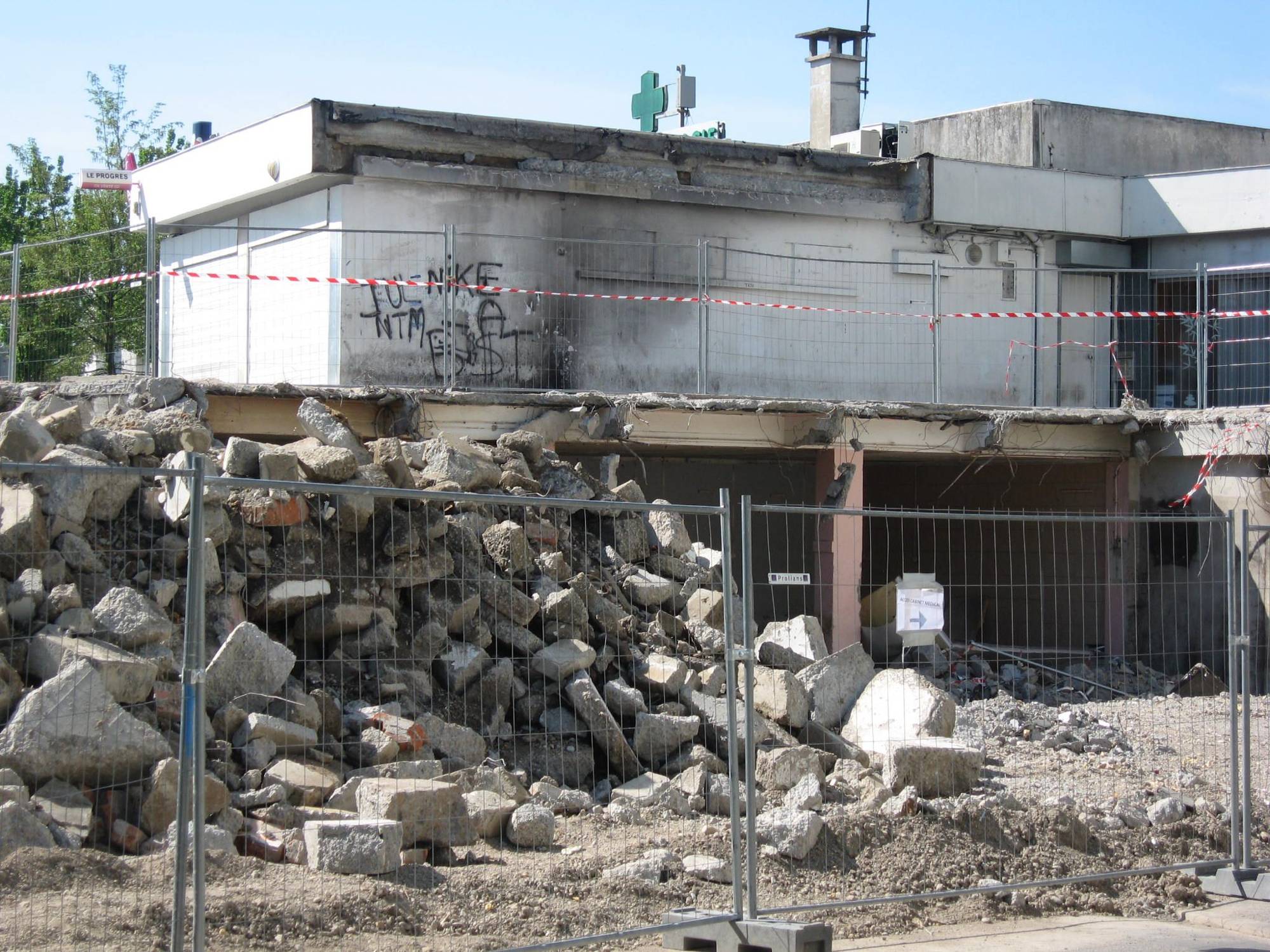 Un bâtiment en cours de démolition, entouré de barrières métalliques et de ruban rouge et blanc, avec un grand tas de gravats au premier plan.