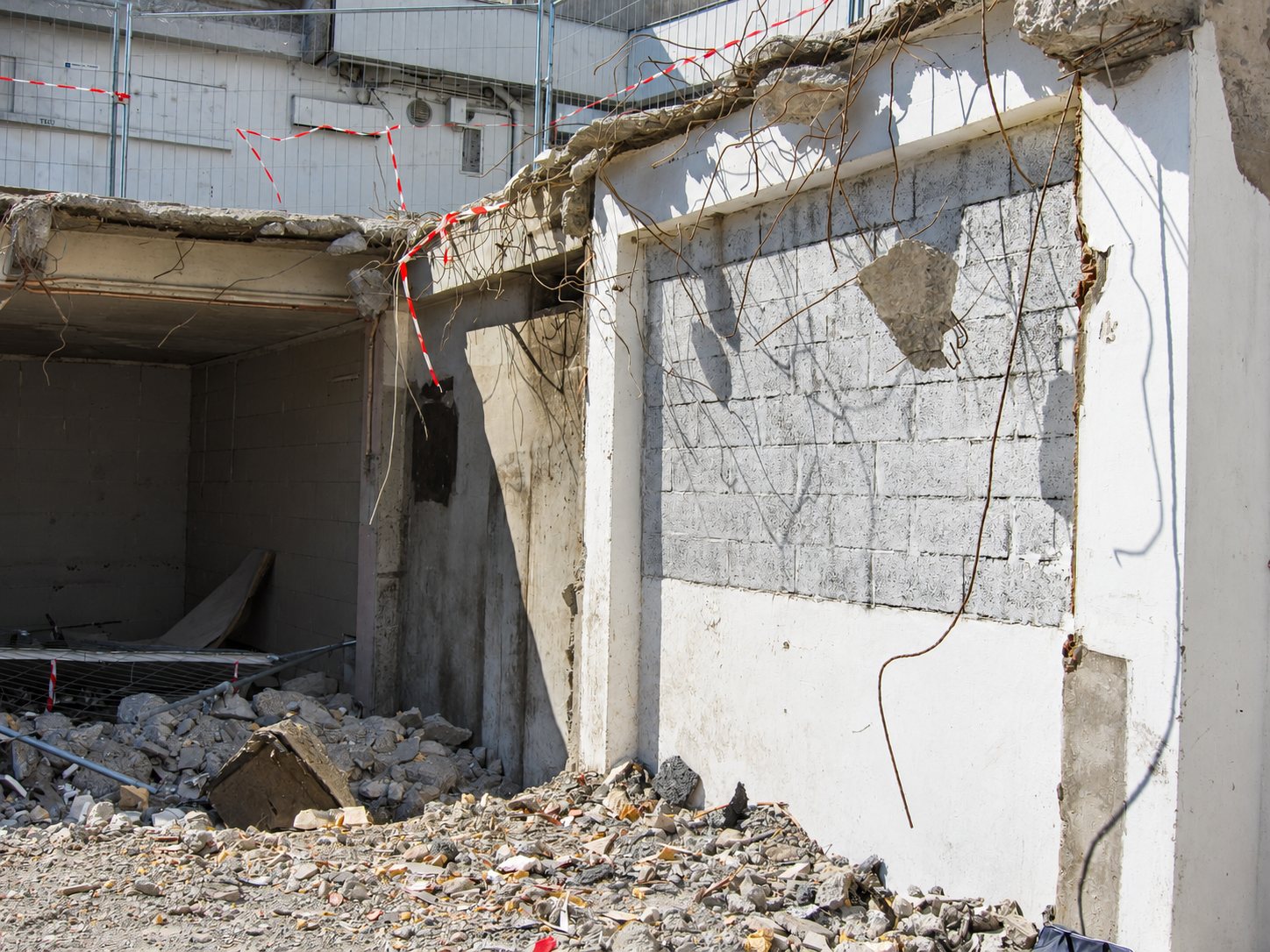 Un chantier de démolition montre un mur en parpaings partiellement détruit, des gravats au sol, des barrières et du ruban de chantier rouge et blanc.