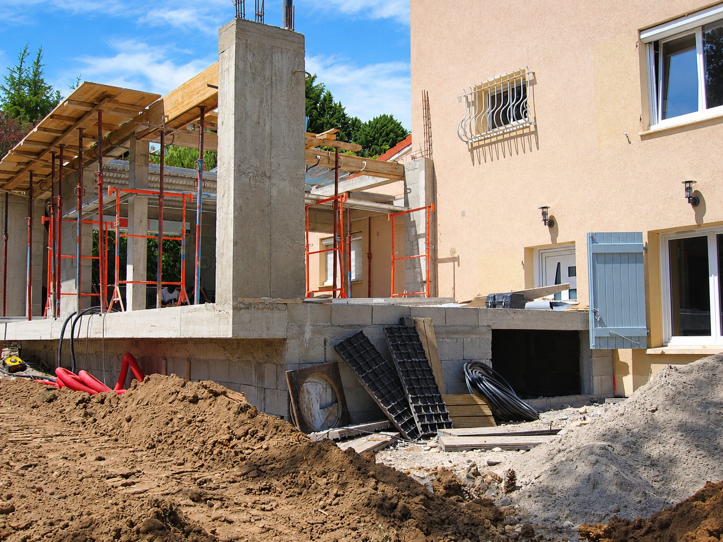 Chantier d'extension d'une maison rose avec un pilier et une dalle en béton soutenus par des étais orange, et des tas de terre au premier plan.