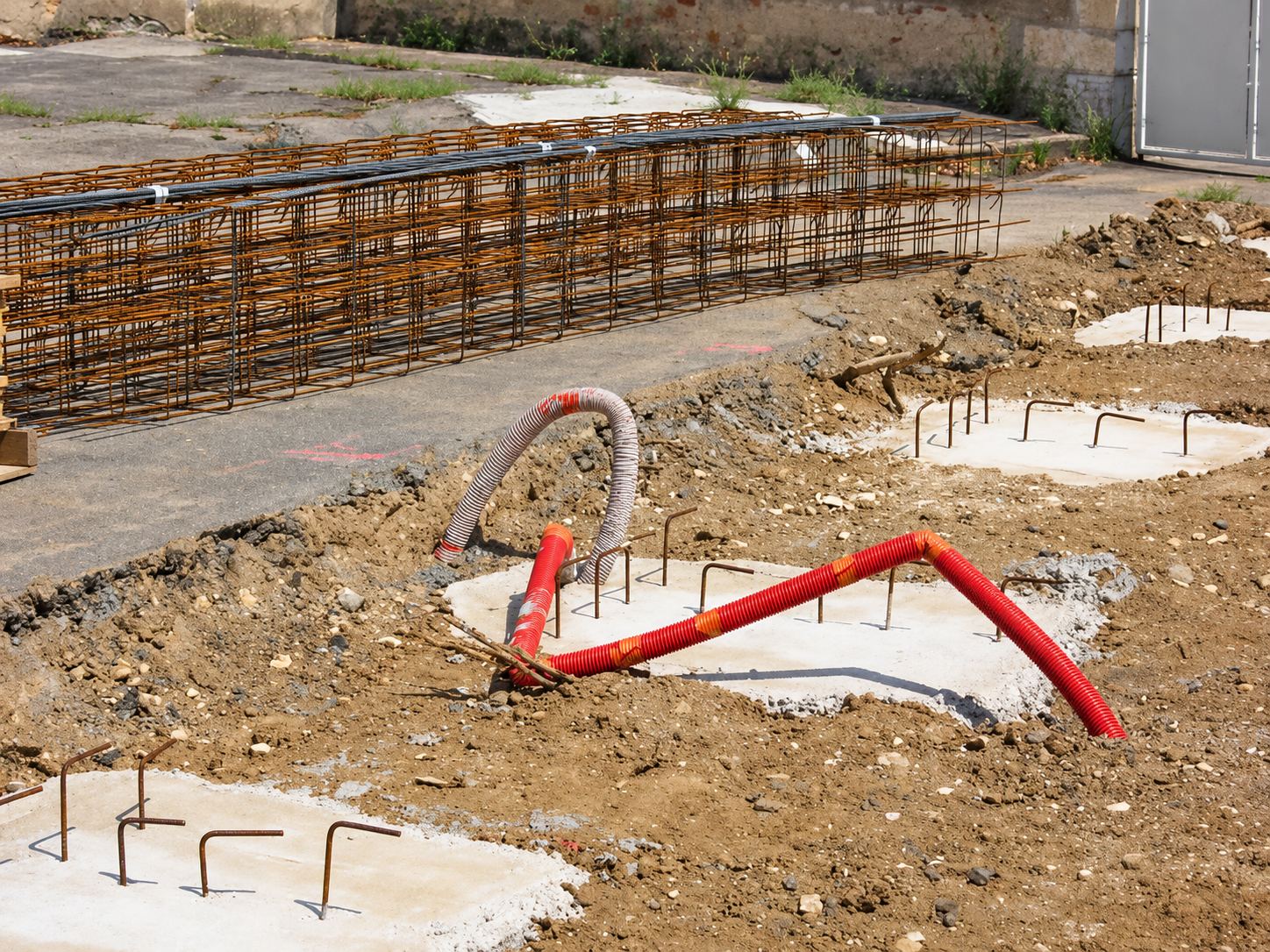 Sur un chantier de terrassement, des plots de béton avec fers en attente et des gaines orange sortent de la terre, devant une grande cage d'armatures métalliques.