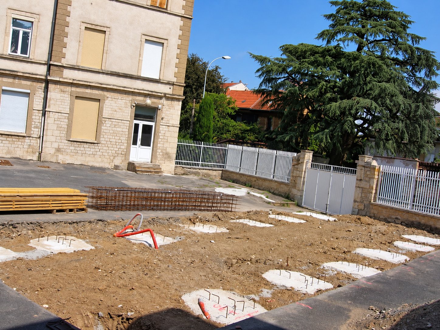 Un chantier de terrassement avec des plots de béton et des armatures en attente devant un bâtiment en pierre et un grand cèdre.