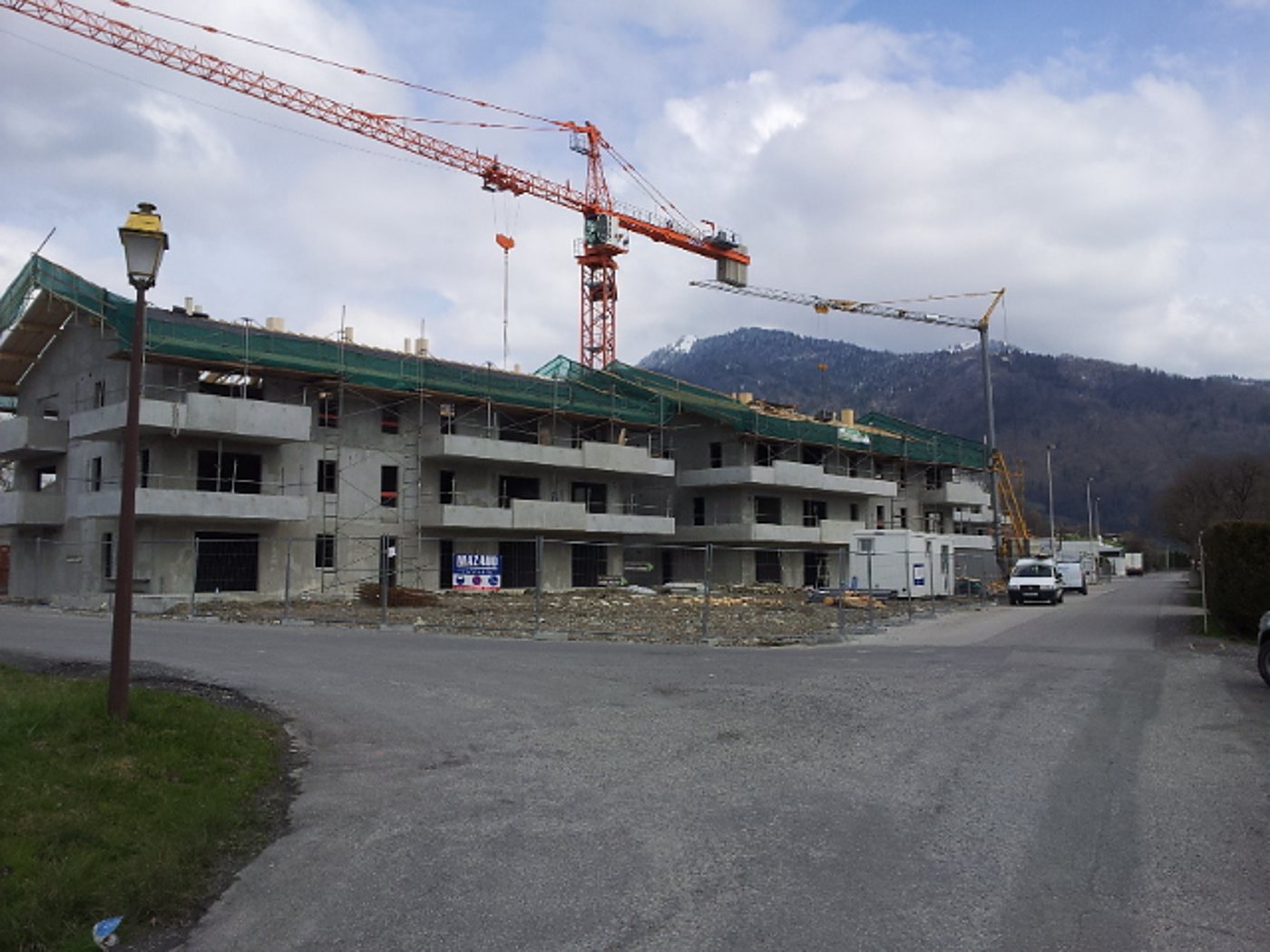 Un immeuble en construction en béton avec des bâches vertes et deux grues, au pied d'une montagne, avec une route et une camionnette au premier plan.