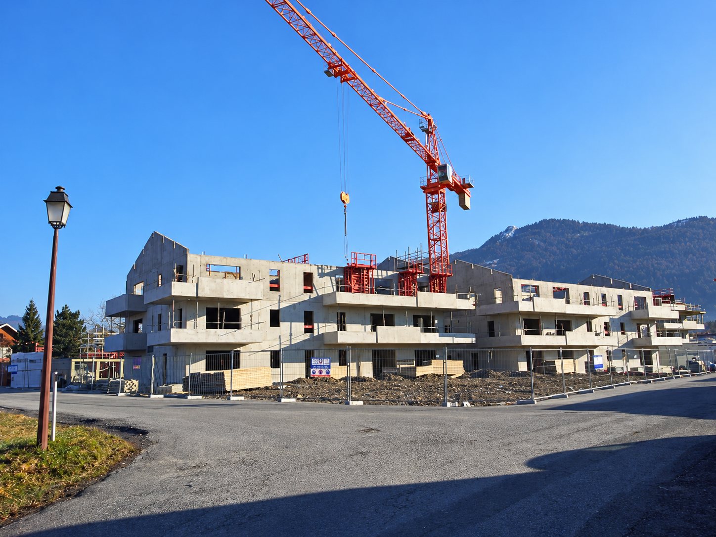 Un immeuble d'habitation en béton en cours de construction, avec une grande grue rouge, au pied d'une montagne sous un ciel bleu.