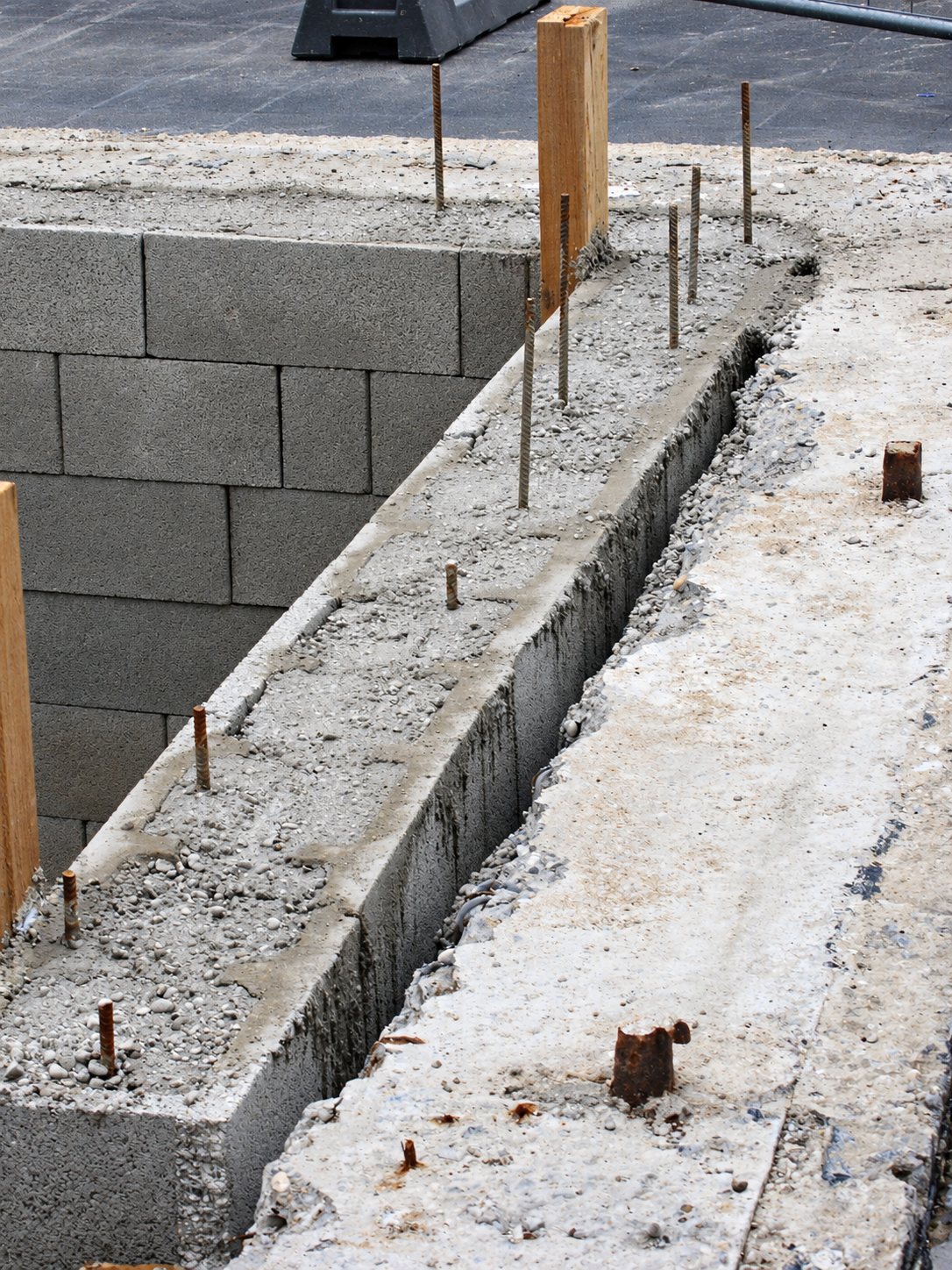 Vue d'un chantier montrant un mur en parpaings, une longrine en béton avec des fers en attente et des planches de bois.