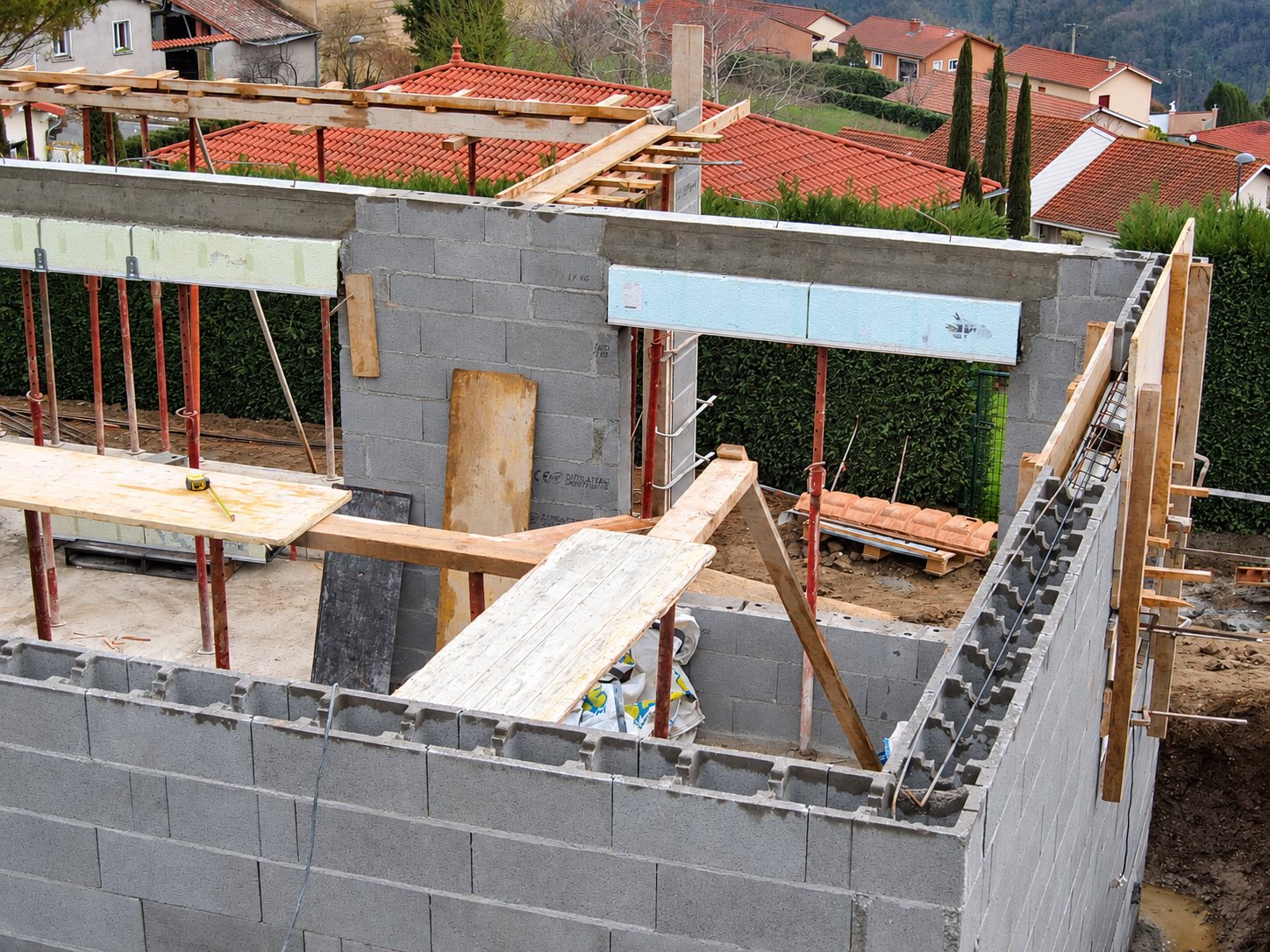 Vue en hauteur d'un chantier de maison en cours avec murs en parpaings, linteaux, étais métalliques et planchers de travail en bois, devant des toits de tuiles rouges.