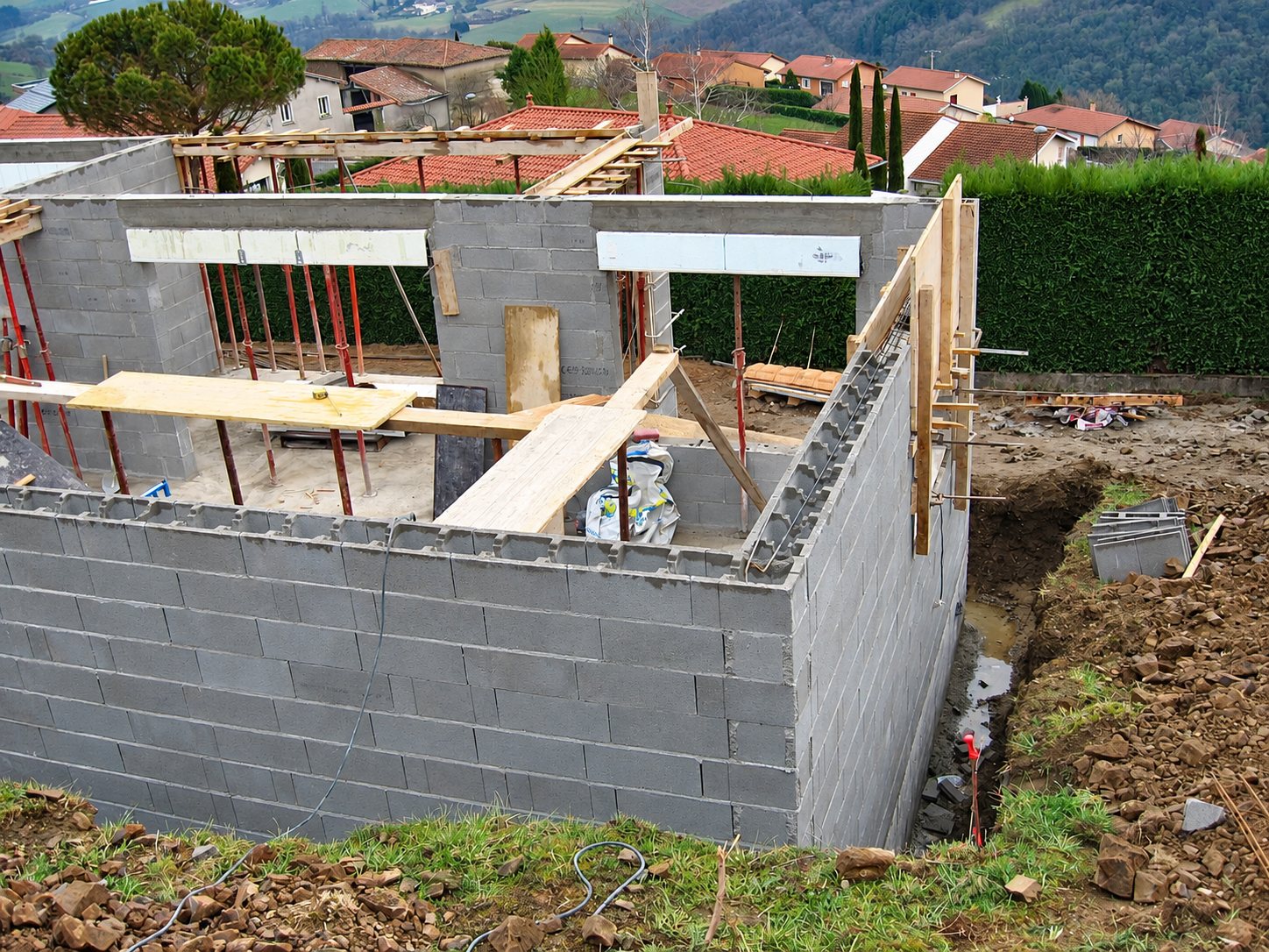 Murs en parpaings d'une maison en construction, avec planches de coffrage et tranchée de terre à droite, dans un paysage vallonné.