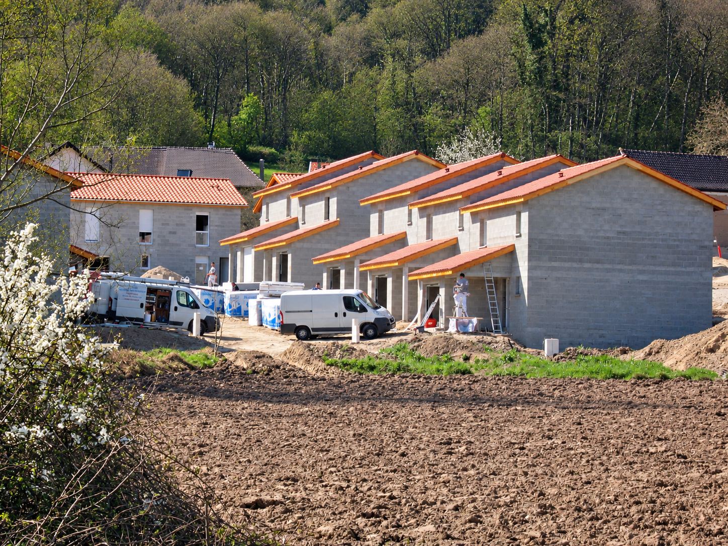 Un lotissement de maisons en parpaings avec toits de tuiles en cours de construction, des camionnettes blanches et des ouvriers sur le chantier au pied d'une colline boisée.