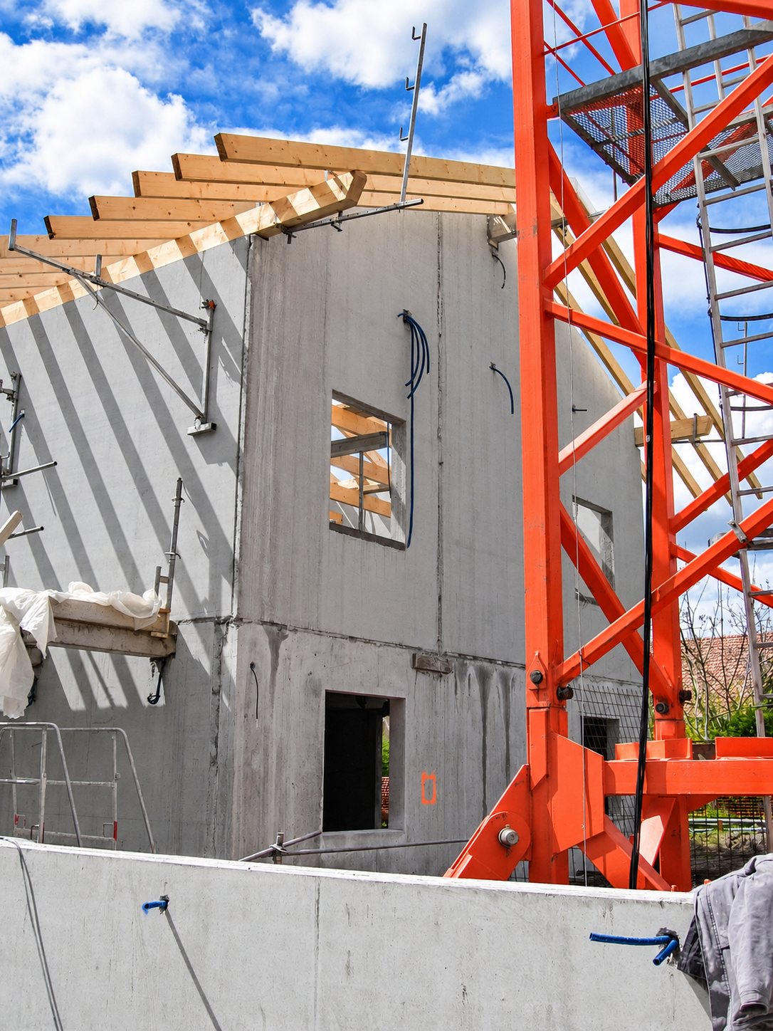 Chantier d'une maison en construction avec murs en béton, début de charpente en bois et grue orange au premier plan.