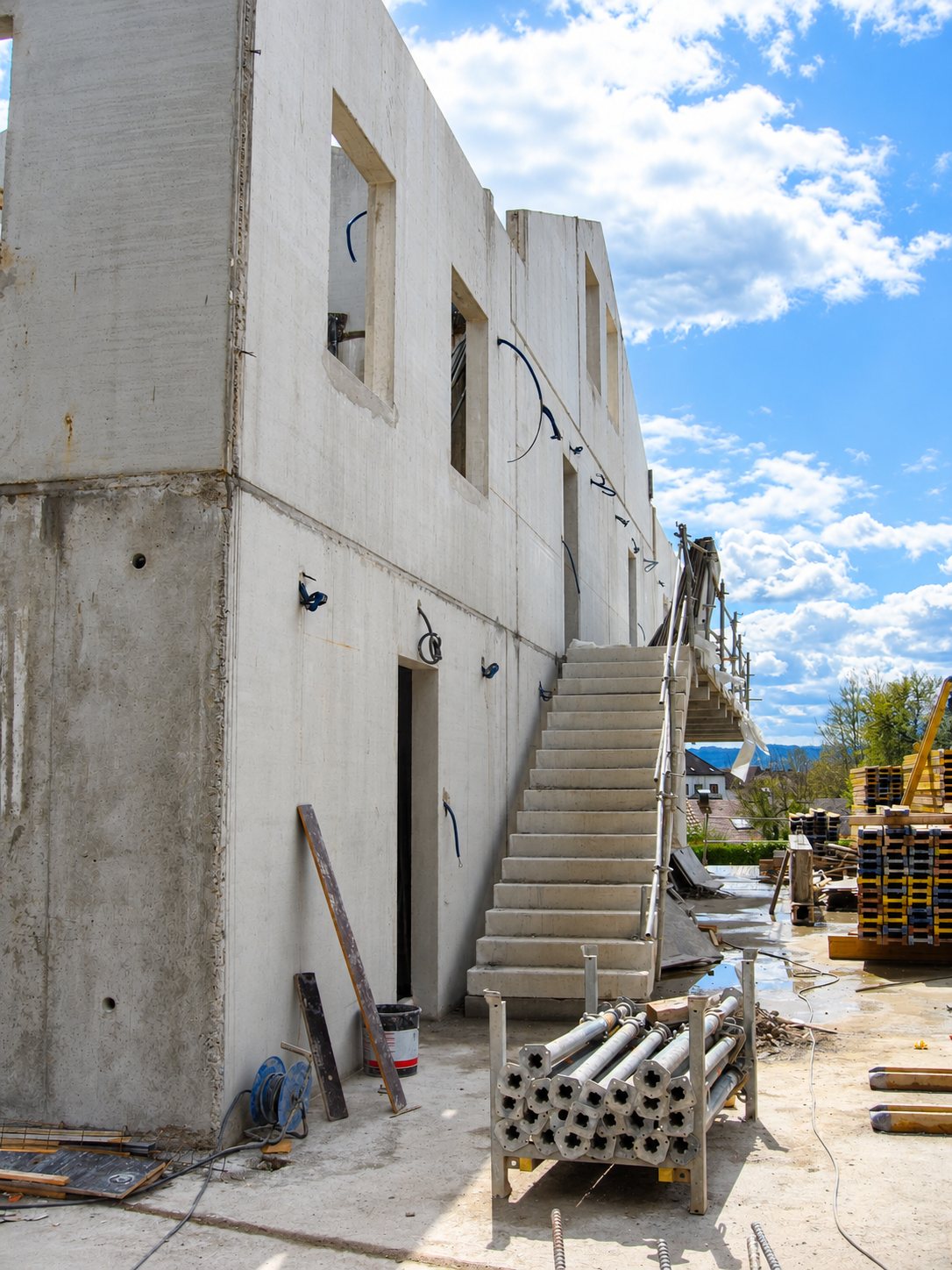 Bâtiment en béton brut en construction avec un escalier extérieur, des étais métalliques rangés sur un support et des planches empilées sous un ciel nuageux.