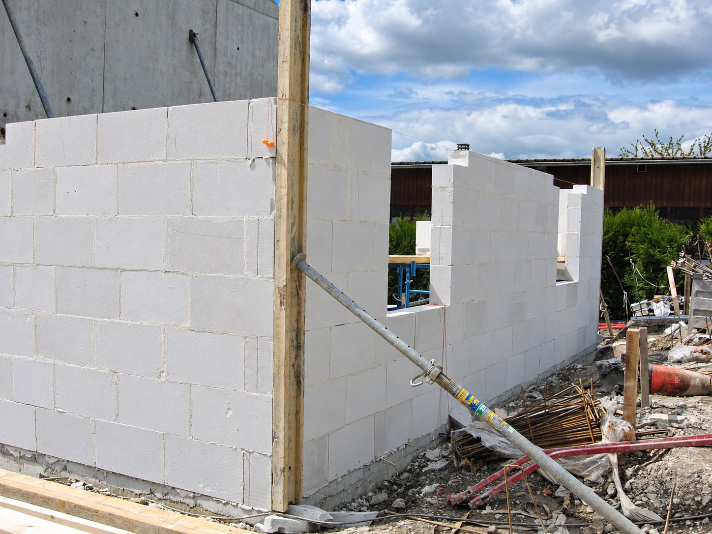 Des murs en blocs de béton cellulaire clair en cours de montage sur un chantier, avec un étai métallique incliné et des gravats au sol sous un ciel nuageux.