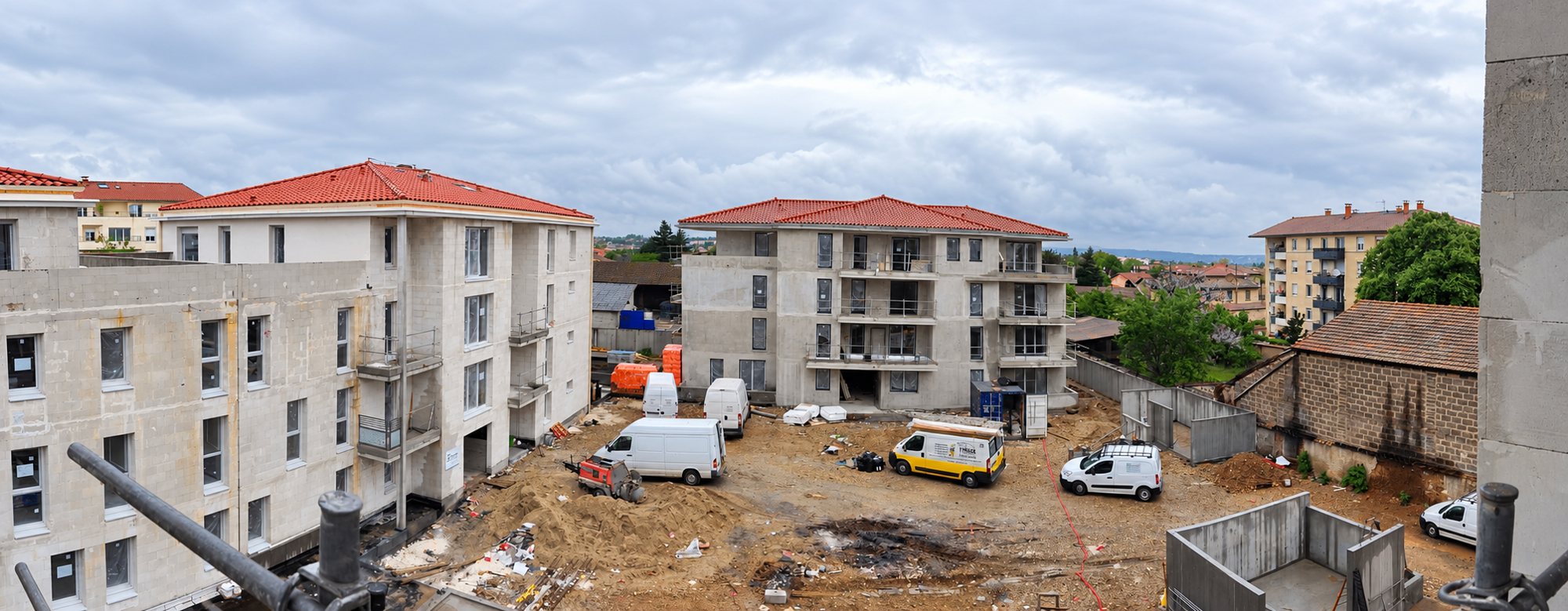 Vue panoramique d'un chantier de résidences en cours de construction, avec des toits de tuiles rouges et des camionnettes garées sur la terre.