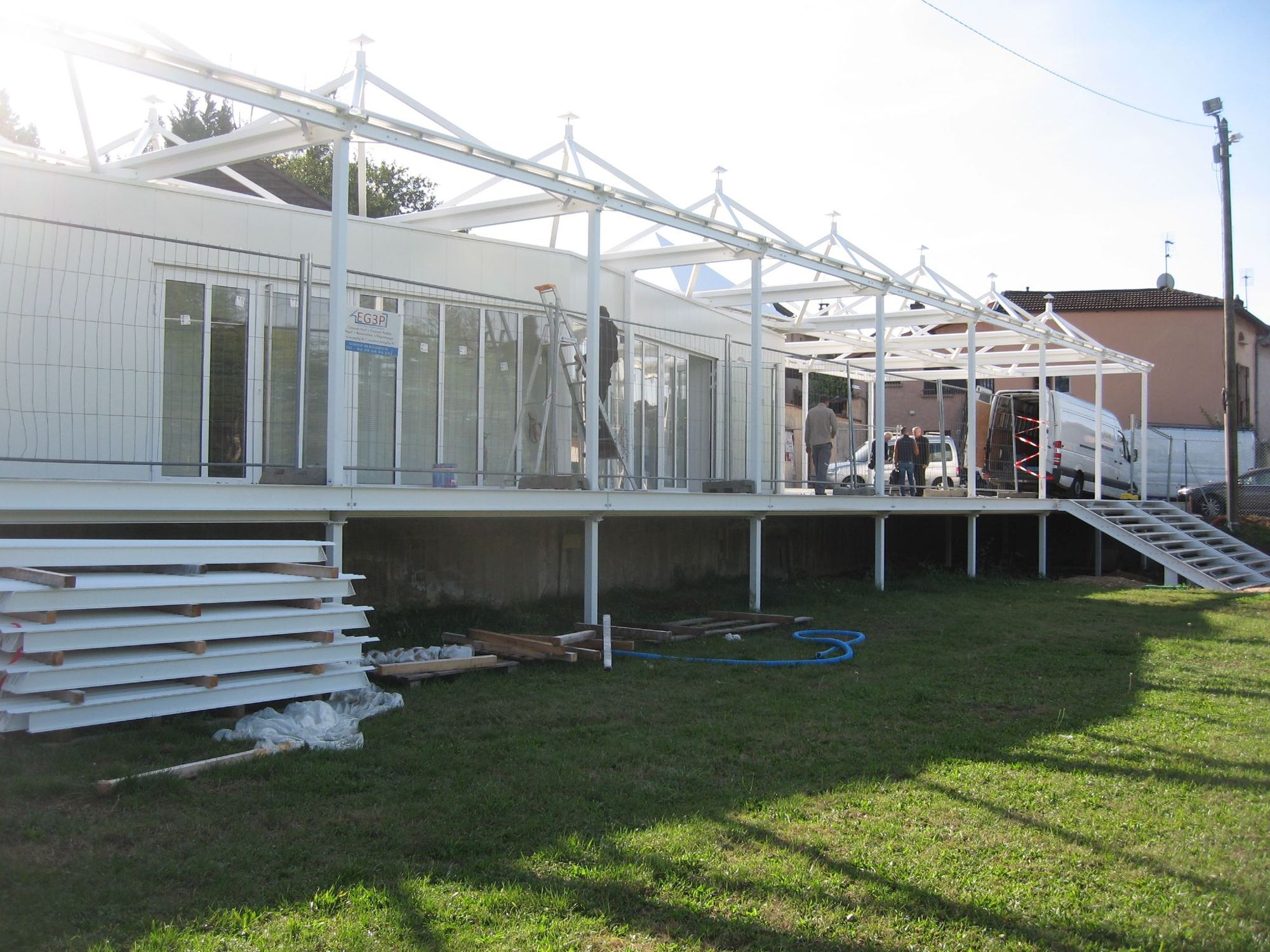 Un chantier de construction montre un bâtiment modulaire blanc surélevé, coiffé d'une structure métallique en cours de montage, avec des ouvriers et une camionnette à proximité.
