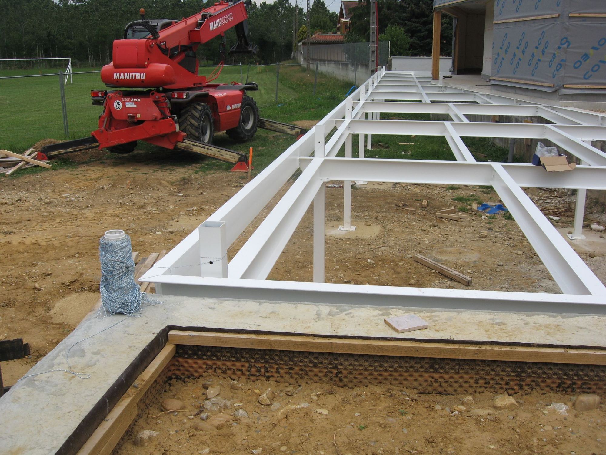 Sur un chantier de terre battue, une ossature en poutres d'acier blanches est posée près d'un chariot télescopique Manitou rouge et d'une maison en construction.