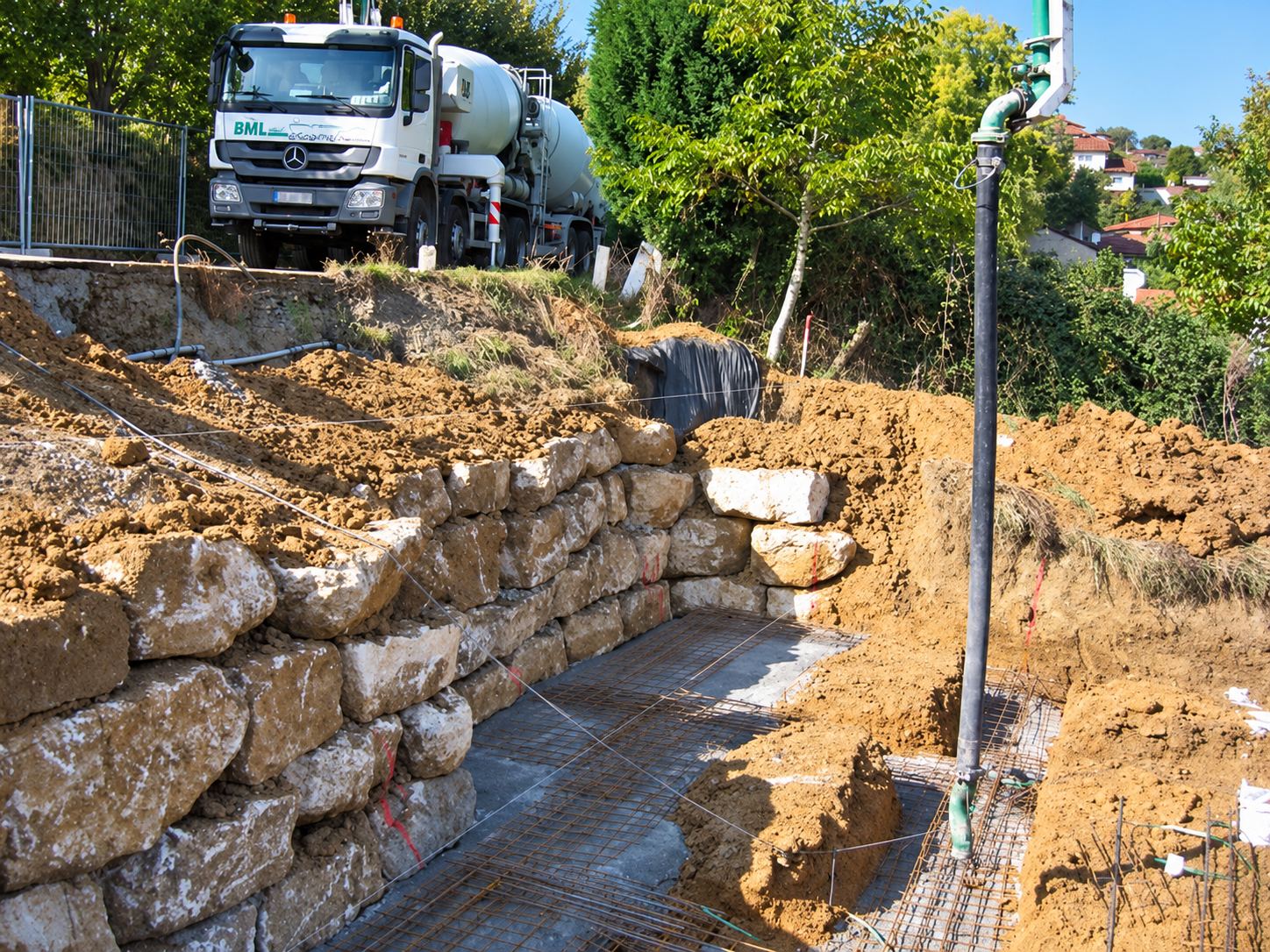 Un chantier de terrassement montre un mur en gros blocs de pierre, un ferraillage au sol et un camion malaxeur Mercedes garé en haut du talus.