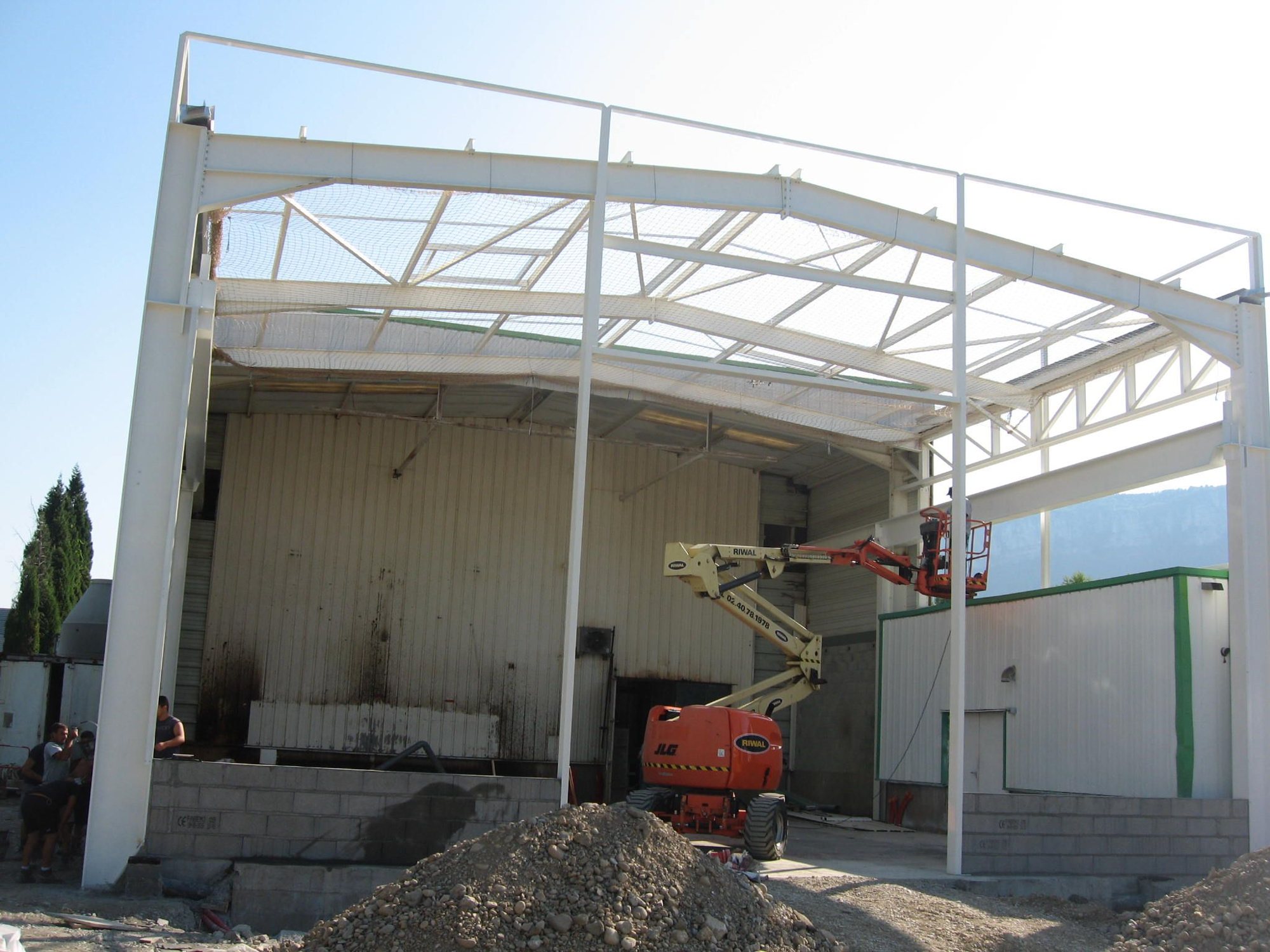 Chantier d'un hangar à charpente métallique blanche, avec une nacelle articulée orange et jaune, un tas de gravats au premier plan et des ouvriers à gauche.