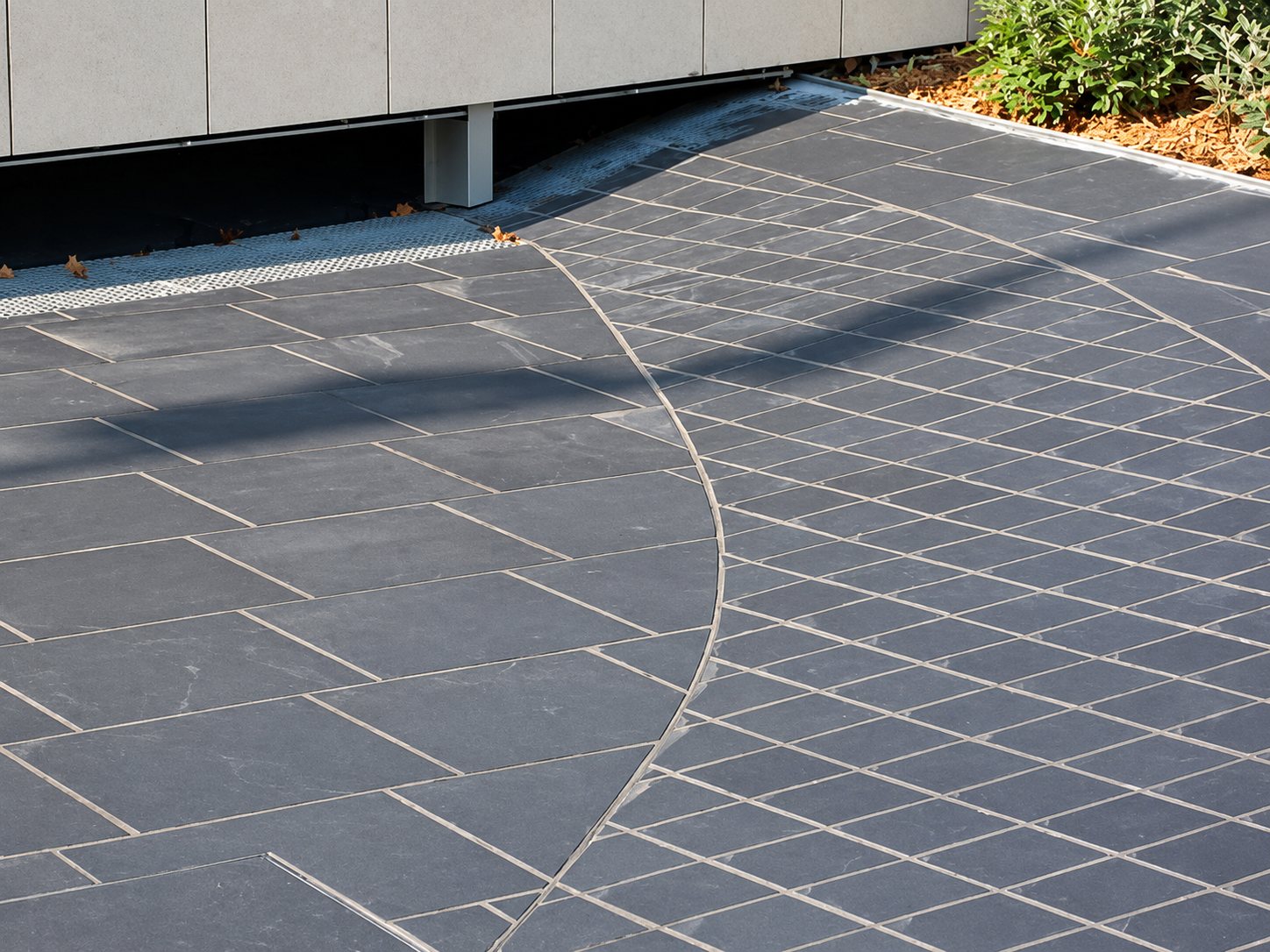 Terrasse extérieure dallée de pierre grise foncée, avec une ligne courbe séparant des grandes dalles rectangulaires et des petits carreaux carrés.