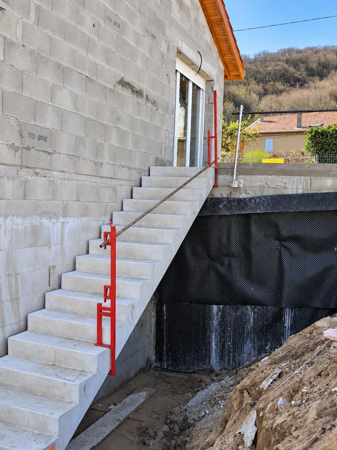 Un escalier extérieur en béton avec rampe provisoire rouge longe le mur en parpaings d'une maison en construction, à côté d'un mur d'étanchéité noir et d'un talus de terre.