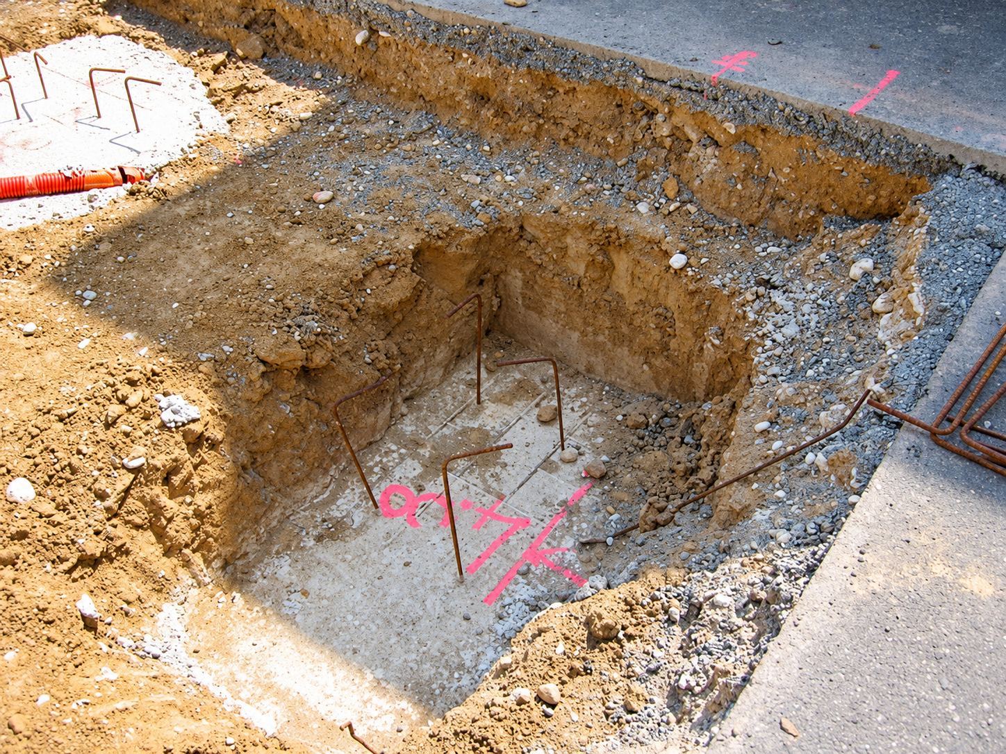 Une fouille carrée creusée dans le sol d'un chantier, avec des fers à béton qui dépassent et des marquages à la peinture rose.