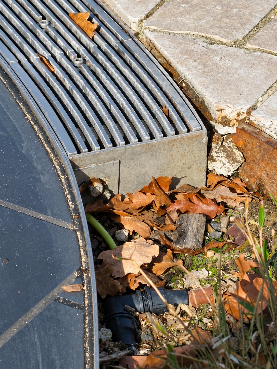 Gros plan sur une grille de caniveau courbe en bordure d'une couverture de bassin, avec des feuilles mortes, un tuyau noir et des dalles de pierre.