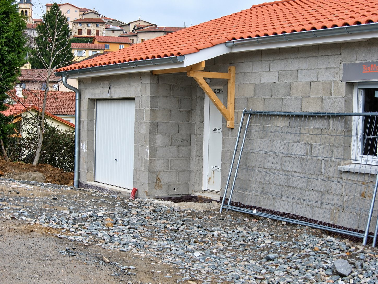 Chantier d'une maison en parpaings apparents avec toit en tuiles oranges, porte de garage blanche, auvent en bois et barrière de chantier.