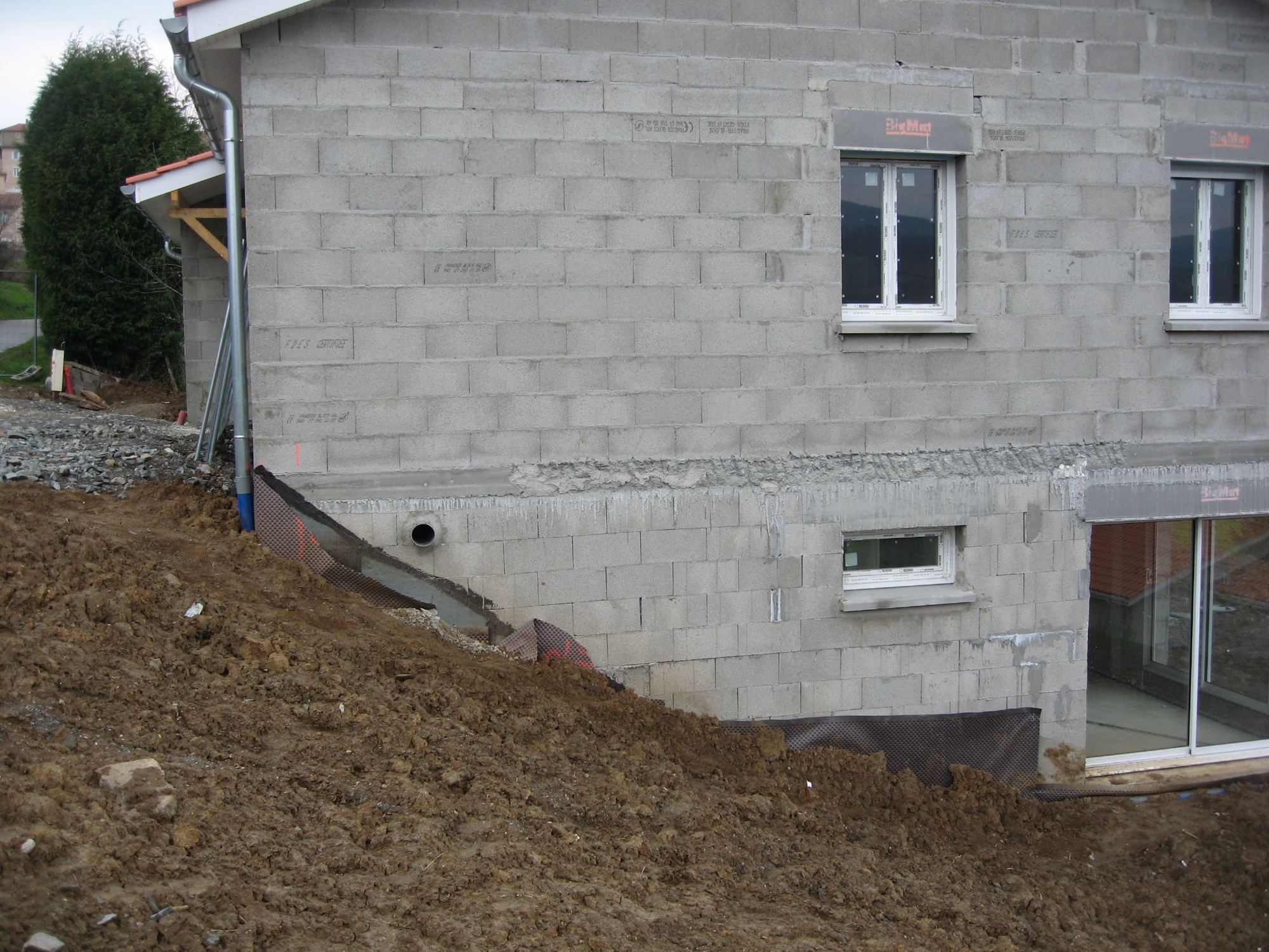 Façade d'une maison en construction en blocs de béton gris, avec fenêtres blanches et talus de terre fraîchement remblayée au premier plan.