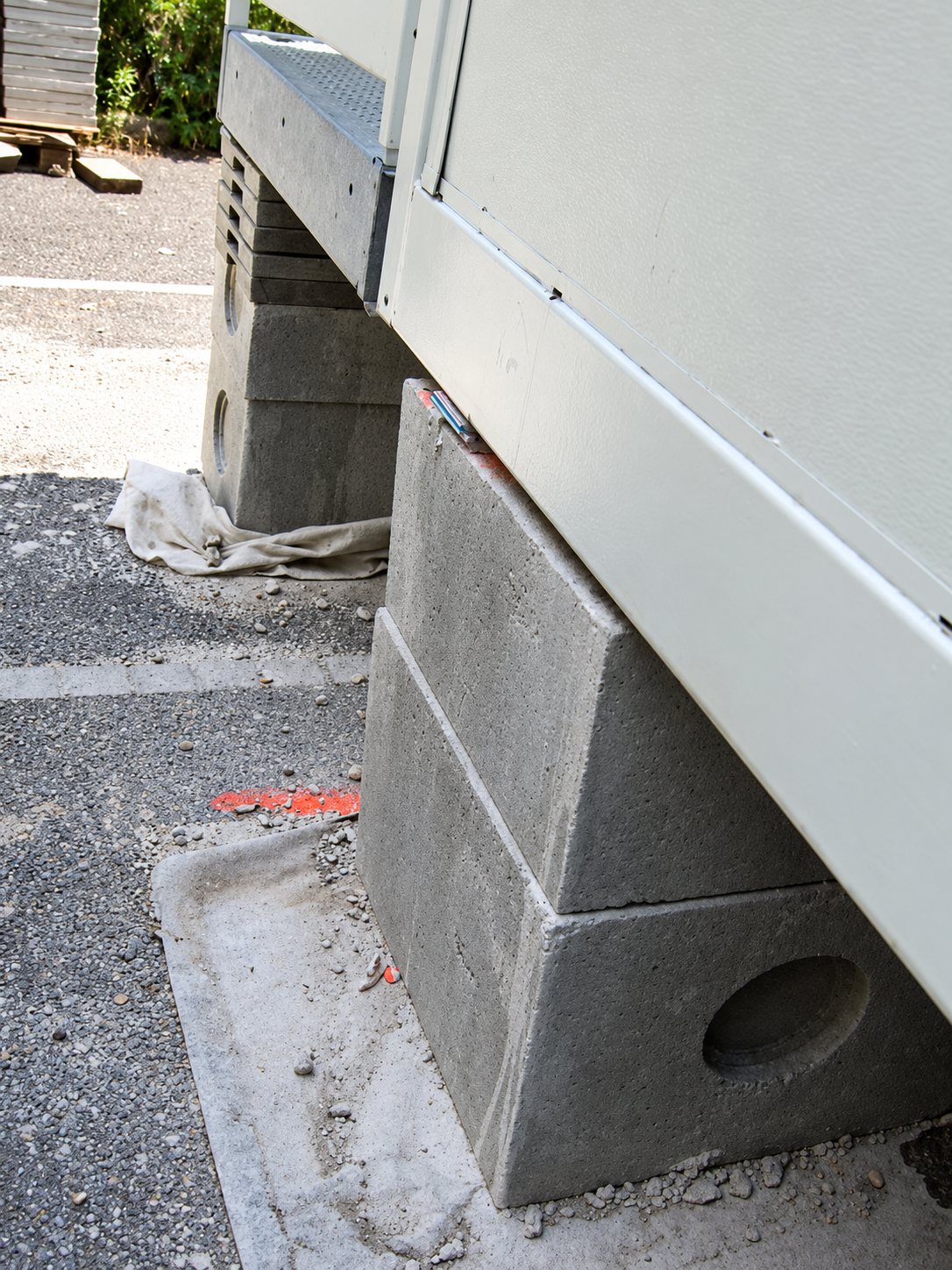 Un module préfabriqué beige repose sur des blocs de béton gris empilés, posés sur un sol gravillonné avec une marque orange peinte.