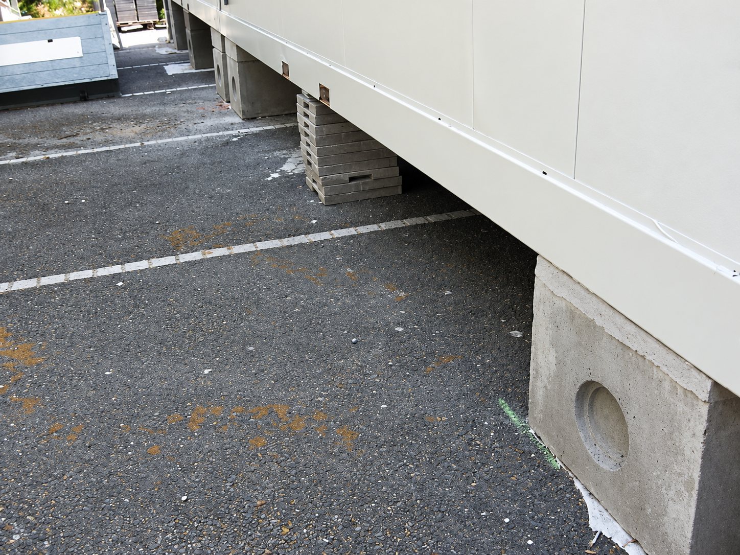 Le bas d'un bâtiment modulaire beige repose sur des plots et des empilements de parpaings en béton, au-dessus d'un sol en enrobé marqué de lignes blanches.
