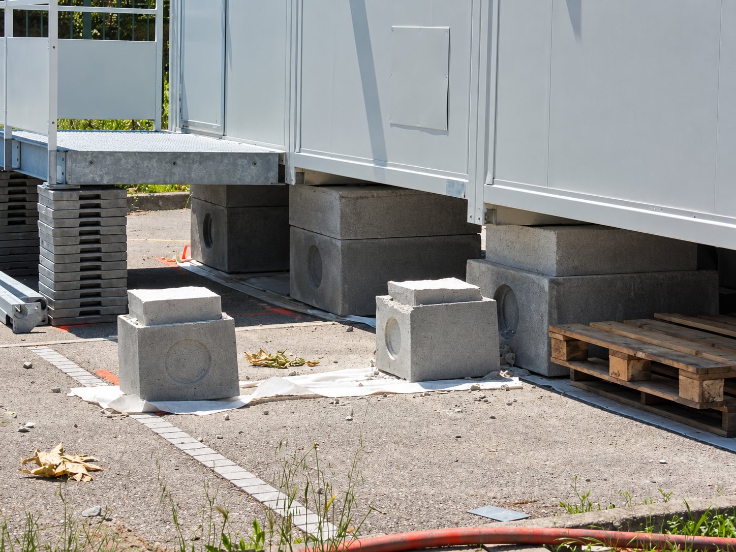 Des bungalows de chantier gris reposent sur des blocs de béton, avec une palette en bois et des plaques de protection posées sur le sol gravillonné.