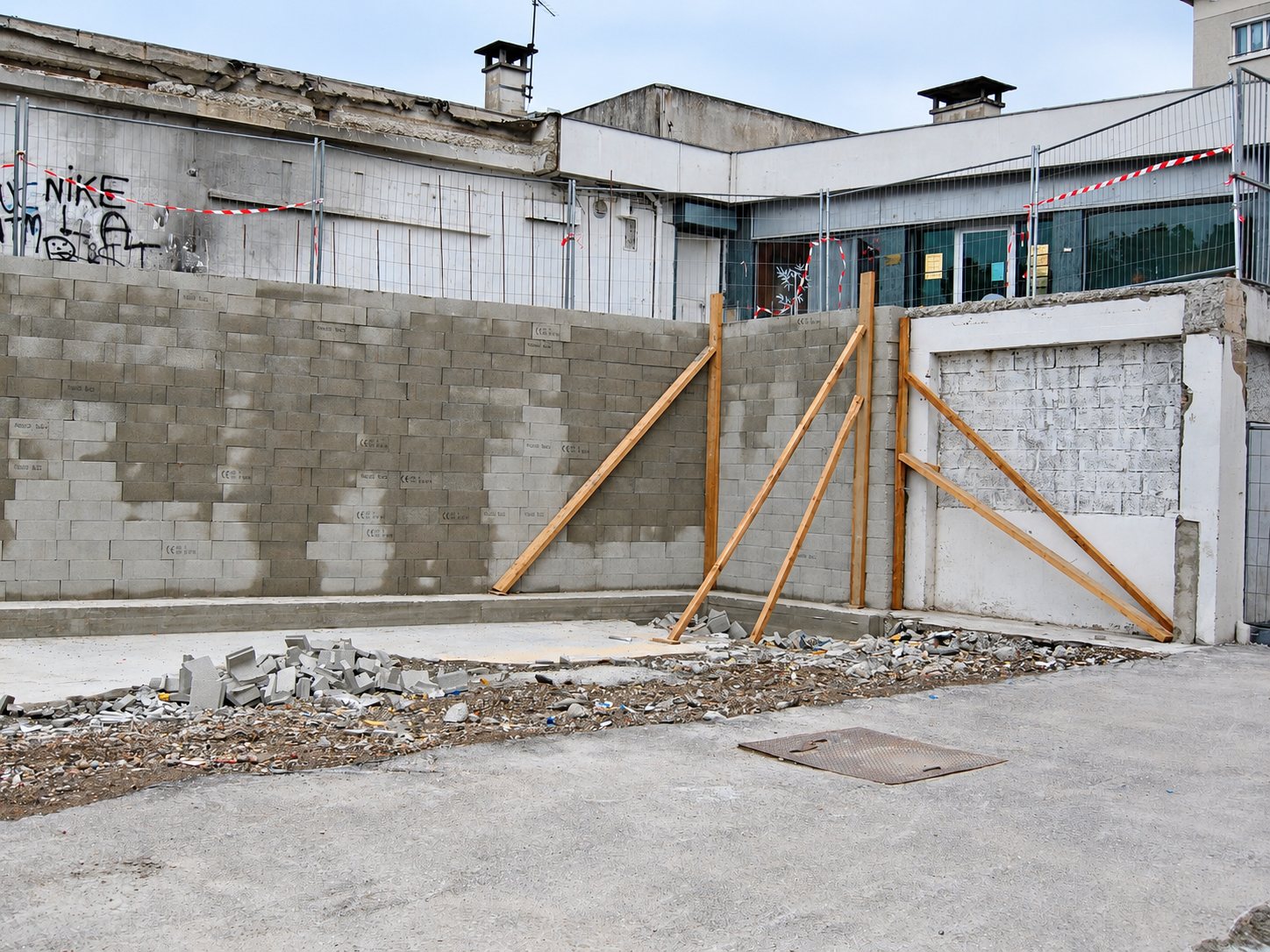 Un mur de parpaings récemment construit est maintenu par des poutres en bois sur un chantier, avec des gravats au sol et des barrières de chantier derrière.