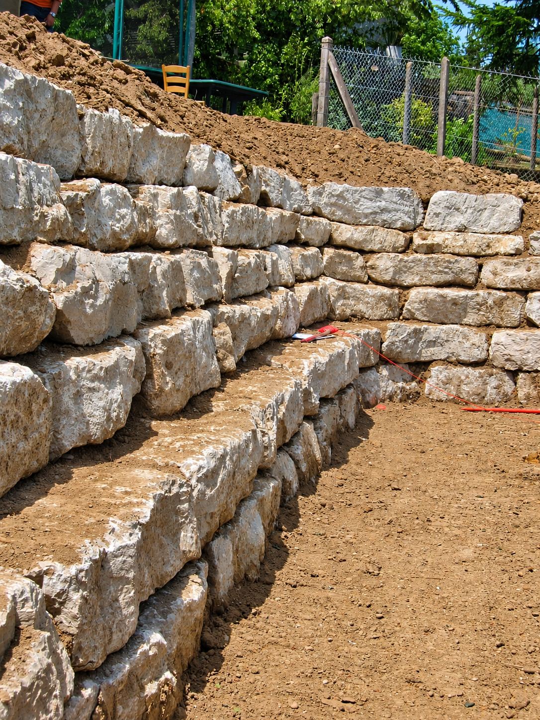 Un mur de soutènement en gros blocs de pierre calcaire empilés en gradins, avec de la terre fraîchement remuée au sommet et un grillage en arrière-plan.