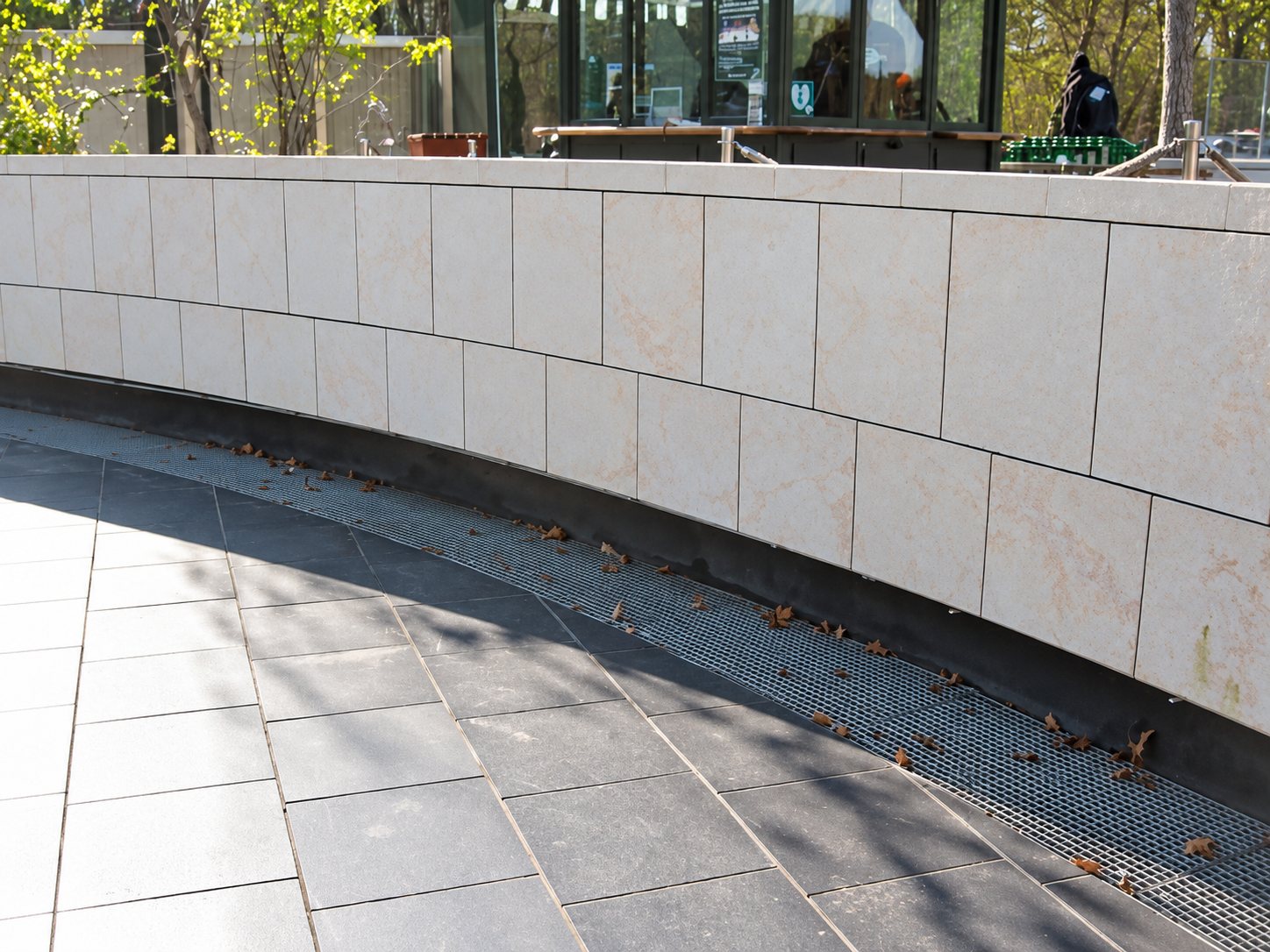 Un muret courbe revêtu de dalles de pierre claire borde un sol dallé gris avec une grille de drainage, devant un kiosque vitré.