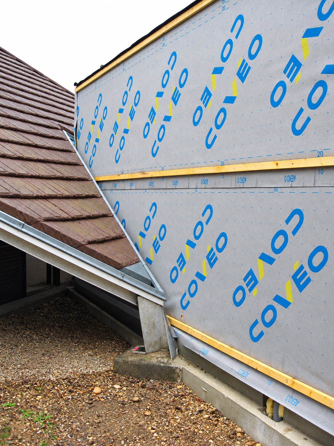 Chantier de toiture montrant une membrane grise imprimée Coveo fixée par des liteaux sur un mur, à côté d'un pan de toit couvert de tuiles brunes.