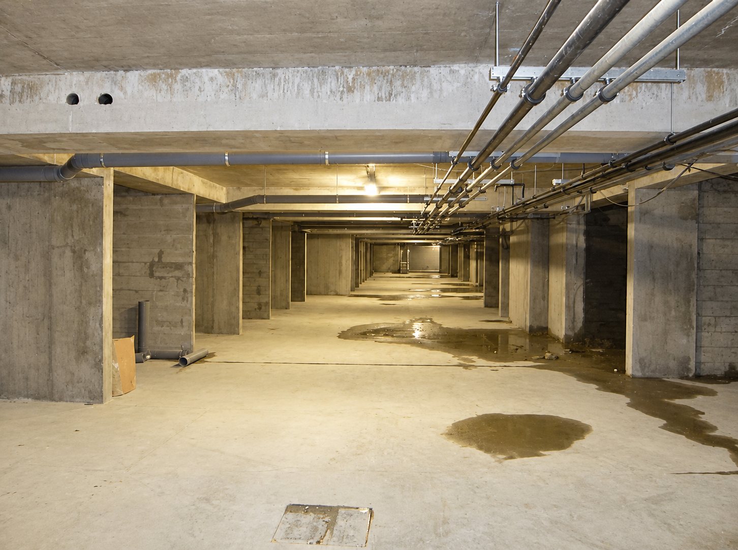 Vaste sous-sol de parking en béton brut avec des tuyaux fixés au plafond, des piliers et des flaques d'eau sur le sol.