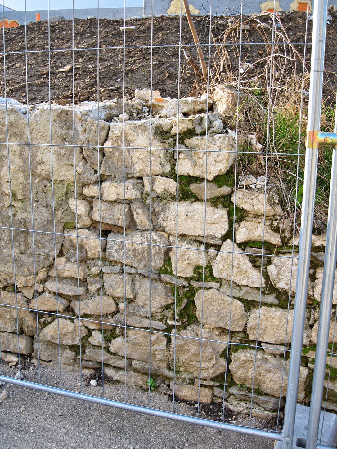Un vieux mur en pierres calcaires partiellement couvert de mousse, protégé par une barrière de chantier métallique, avec de la terre remuée au-dessus.